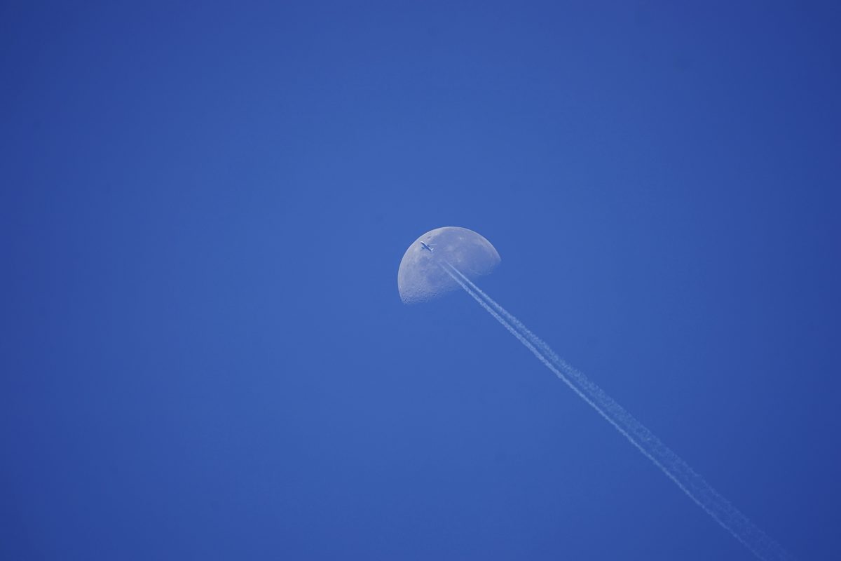 Airliner on the upper edge of the daytime moon, twin contrails streaming away across the disc