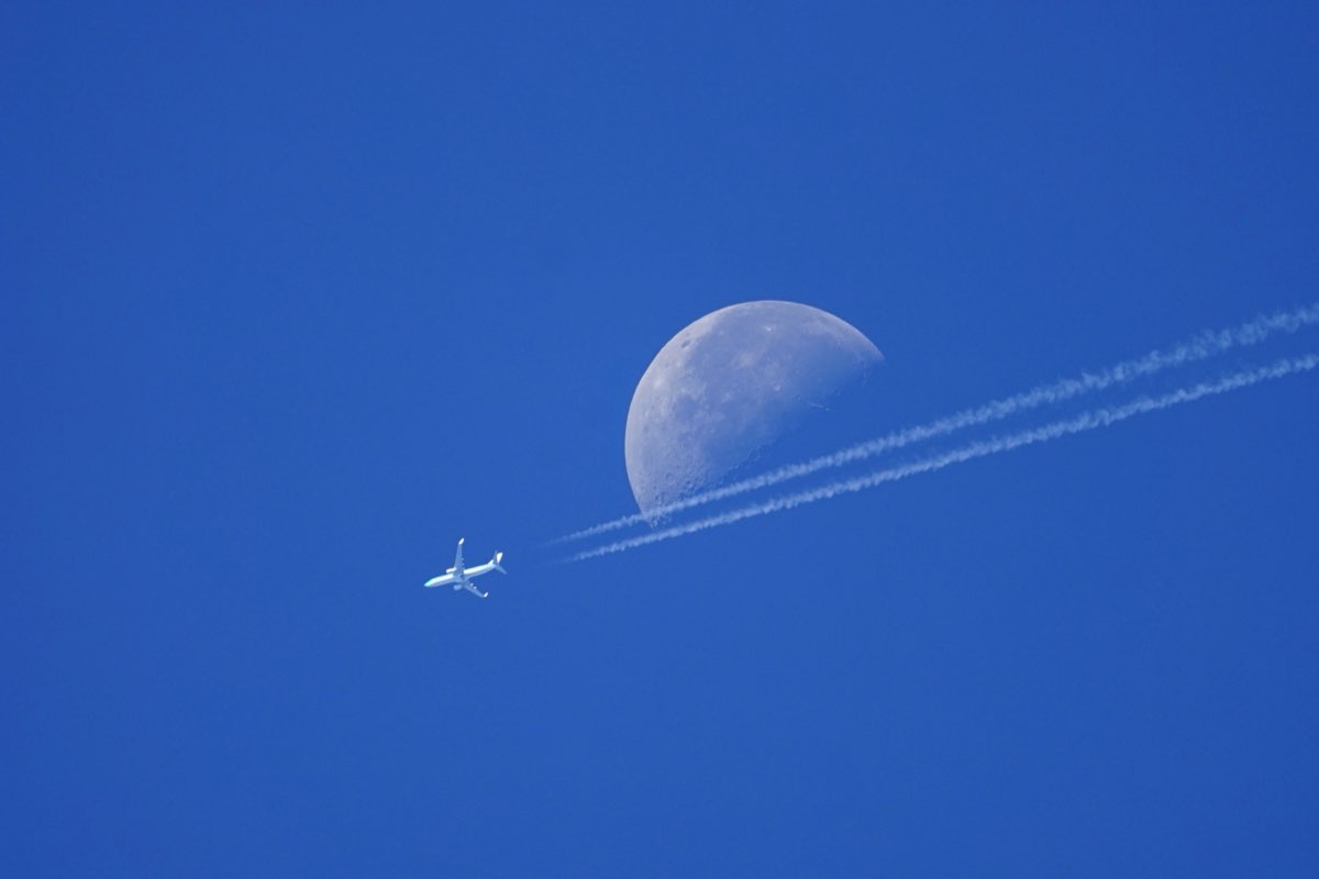 Airliner just past the daytime moon, its contrails cutting across the moon&rsquo;s lower edge