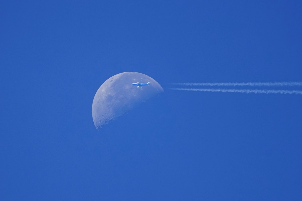Blue-tailed airliner on the right edge of the daytime moon, contrails trailing off to the right