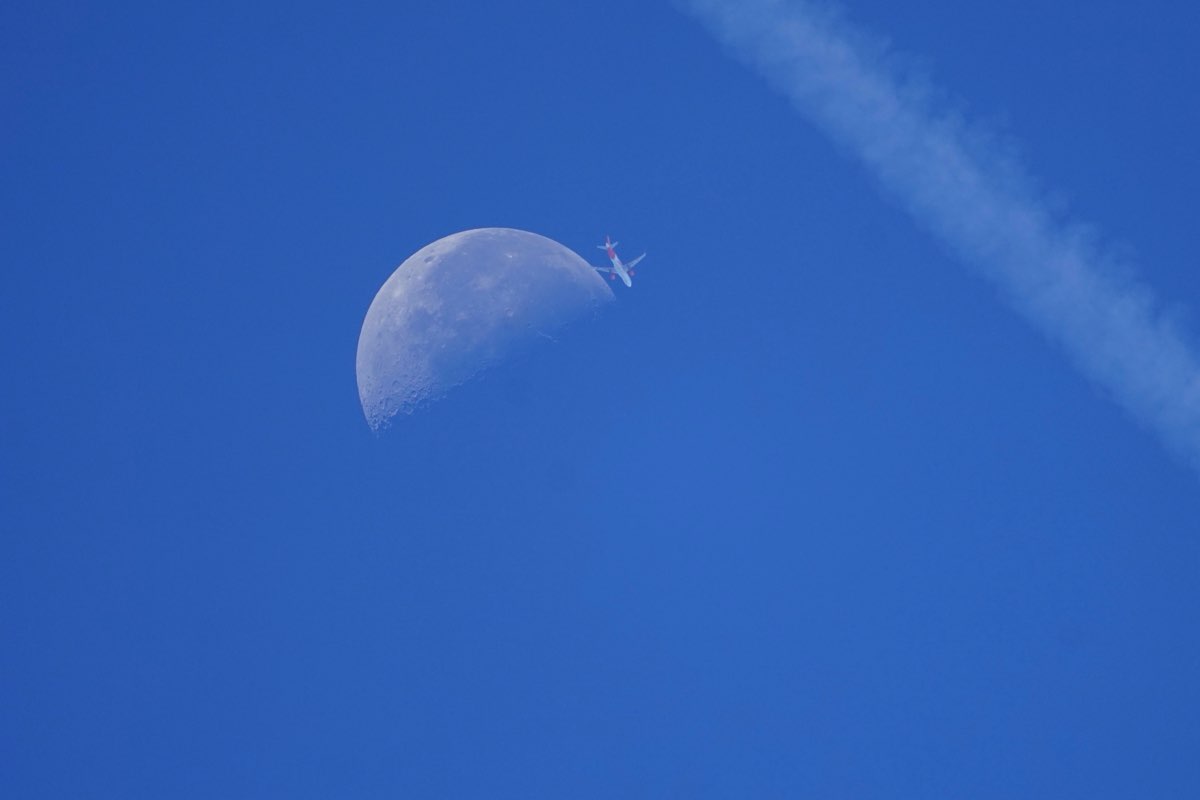 Airliner at the upper-right edge of the daytime moon, an older contrail sweeping across the sky