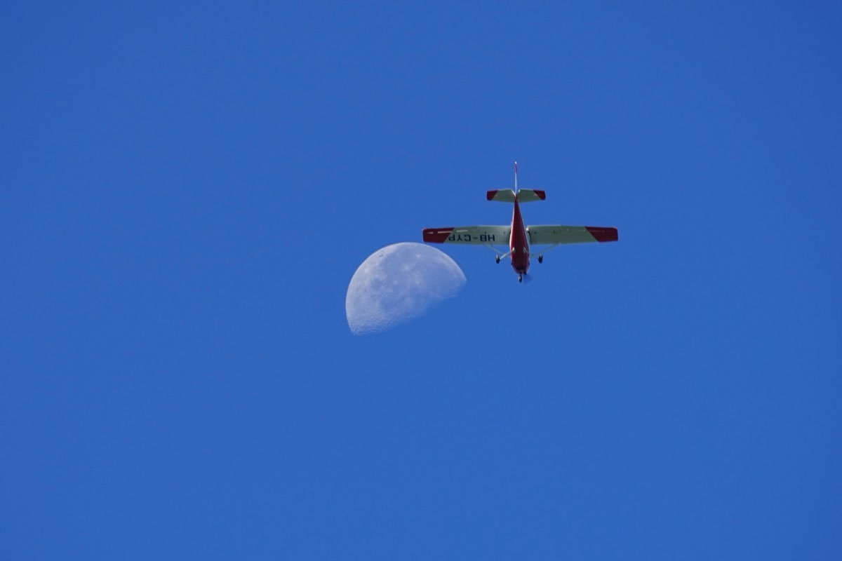Red-and-white Cessna 152 seen from below, passing just to the right of the daytime moon