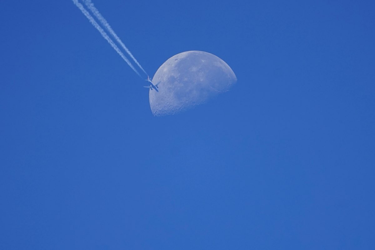 Airliner on the lower-left edge of the daytime moon, twin contrails trailing away to the upper left