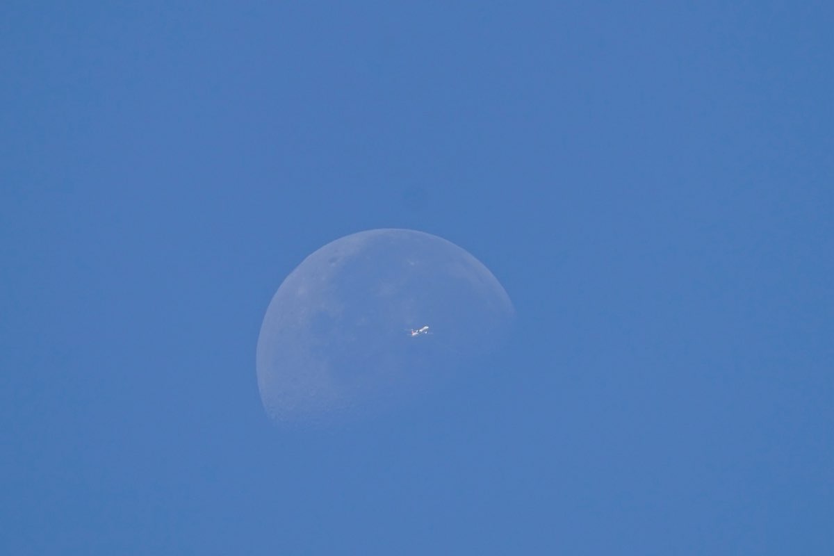 An airliner caught mid-transit, silhouetted directly against the face of the daytime moon