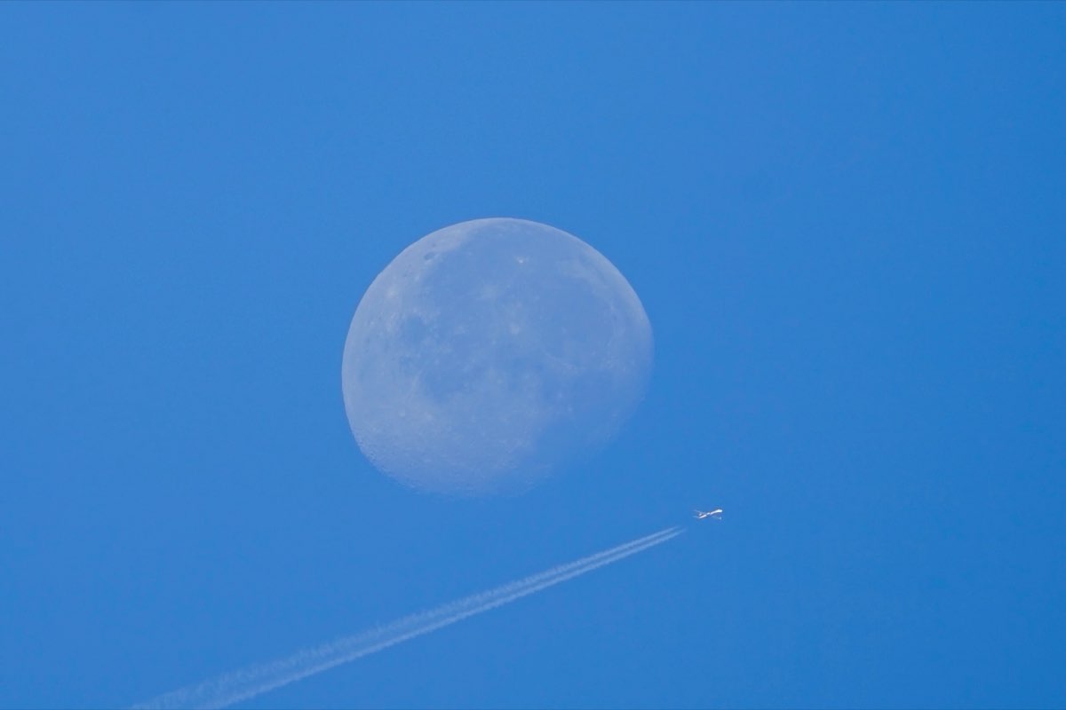 An airliner and its contrail passing just below the daytime moon